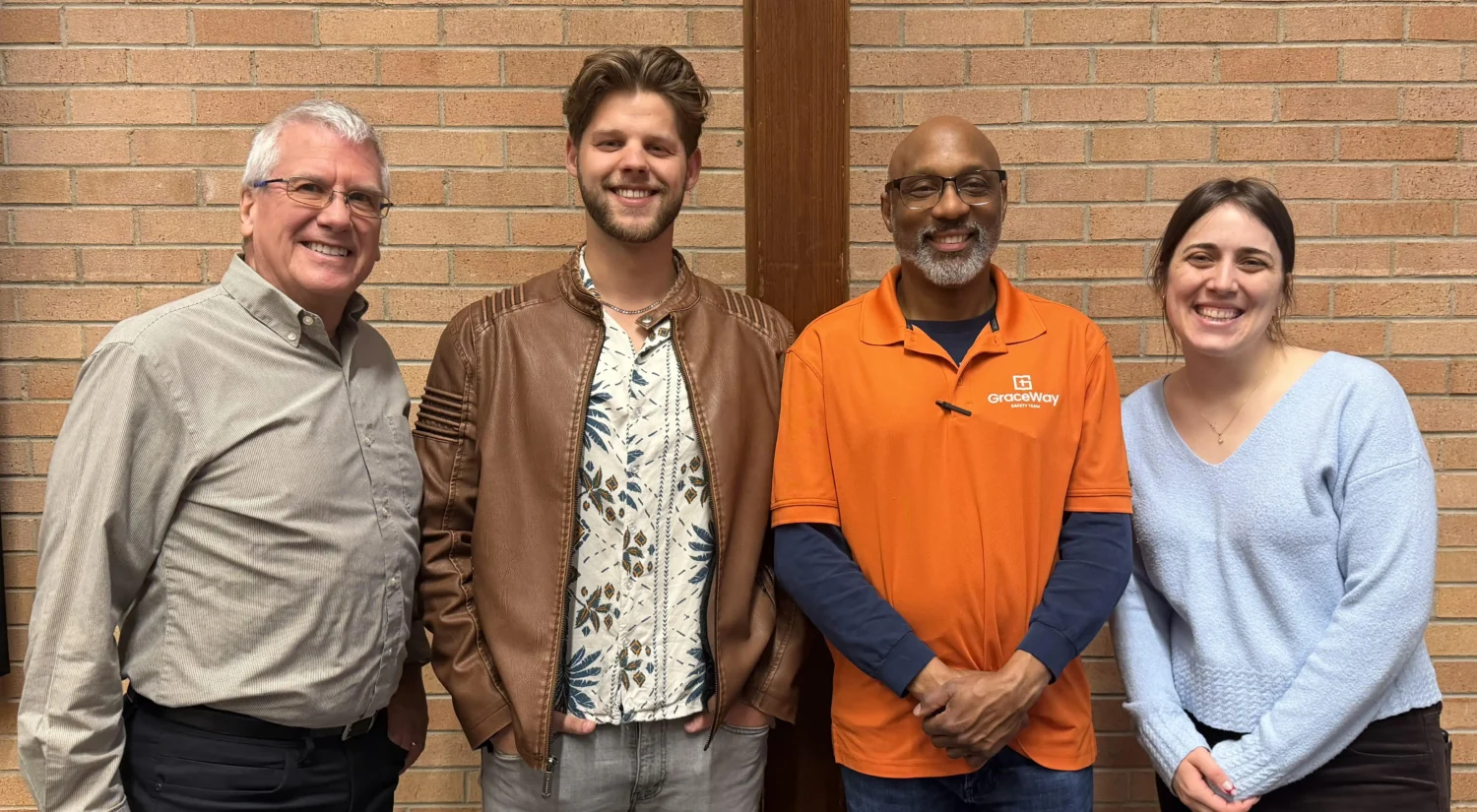 Group of four individuals smiling together, wearing casual attire, with one person in an orange GraceWay shirt, standing in front of a brick wall, representing the GraceWay Bible Church mission team involved in outreach initiatives.